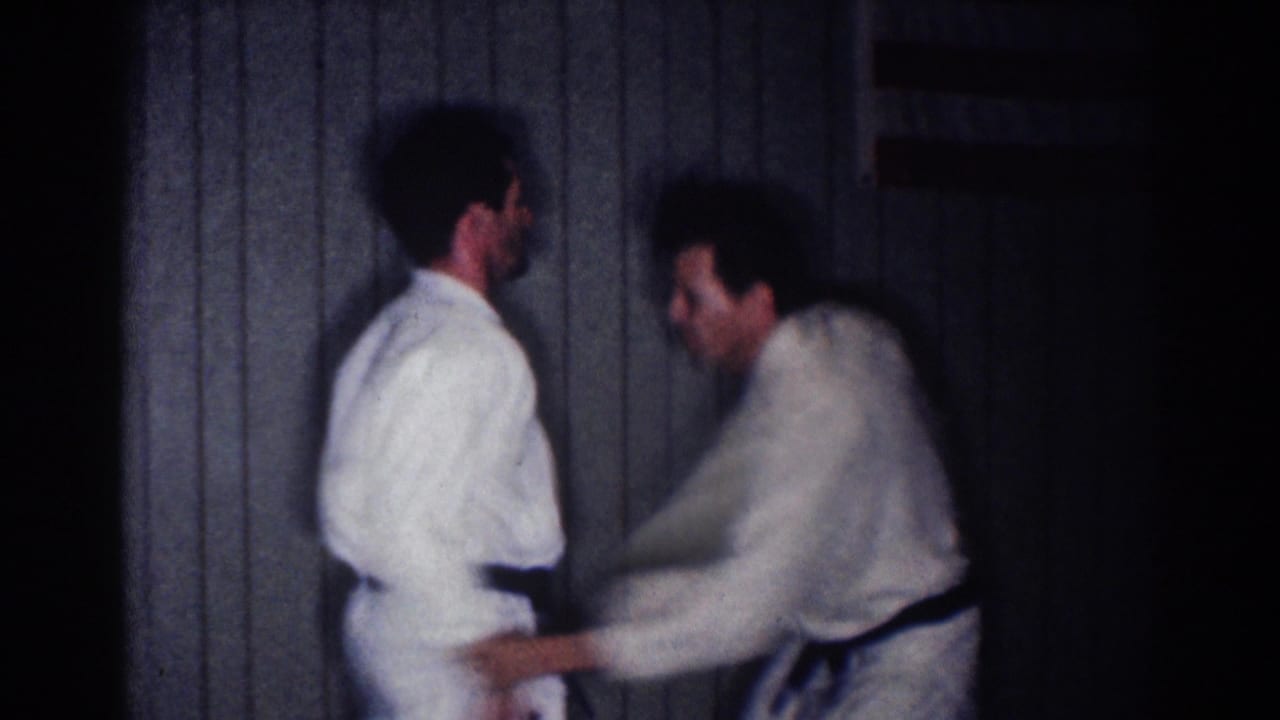 Two adult men in white gi and black belts practice a close defensive movement indoors in the 1967 Los Angeles home-movie clip.