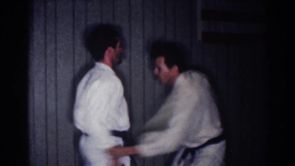 Two adult men in white gi and black belts practice a close defensive movement indoors in the 1967 Los Angeles home-movie clip.
