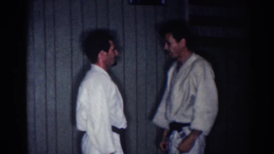 Two adult men in white martial arts uniforms face one another during a pause or reset in the supported 1967 Los Angeles footage.