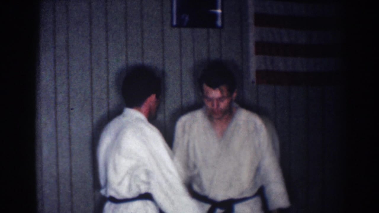 Two adult men continue a karate practice exchange indoors, with a small flag-like object visible above them in the 1967 Los Angeles home movie.