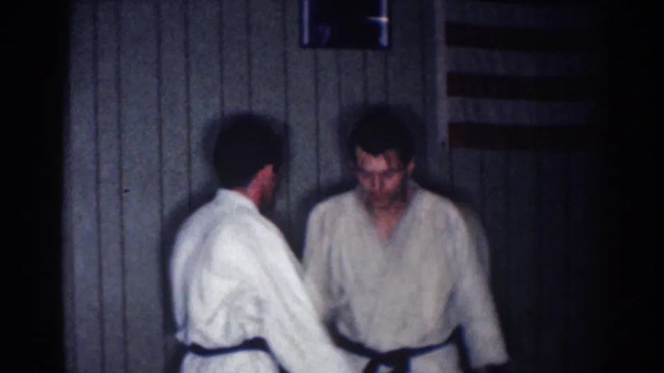 Two adult men continue a karate practice exchange indoors, with a small flag-like object visible above them in the 1967 Los Angeles home movie.
