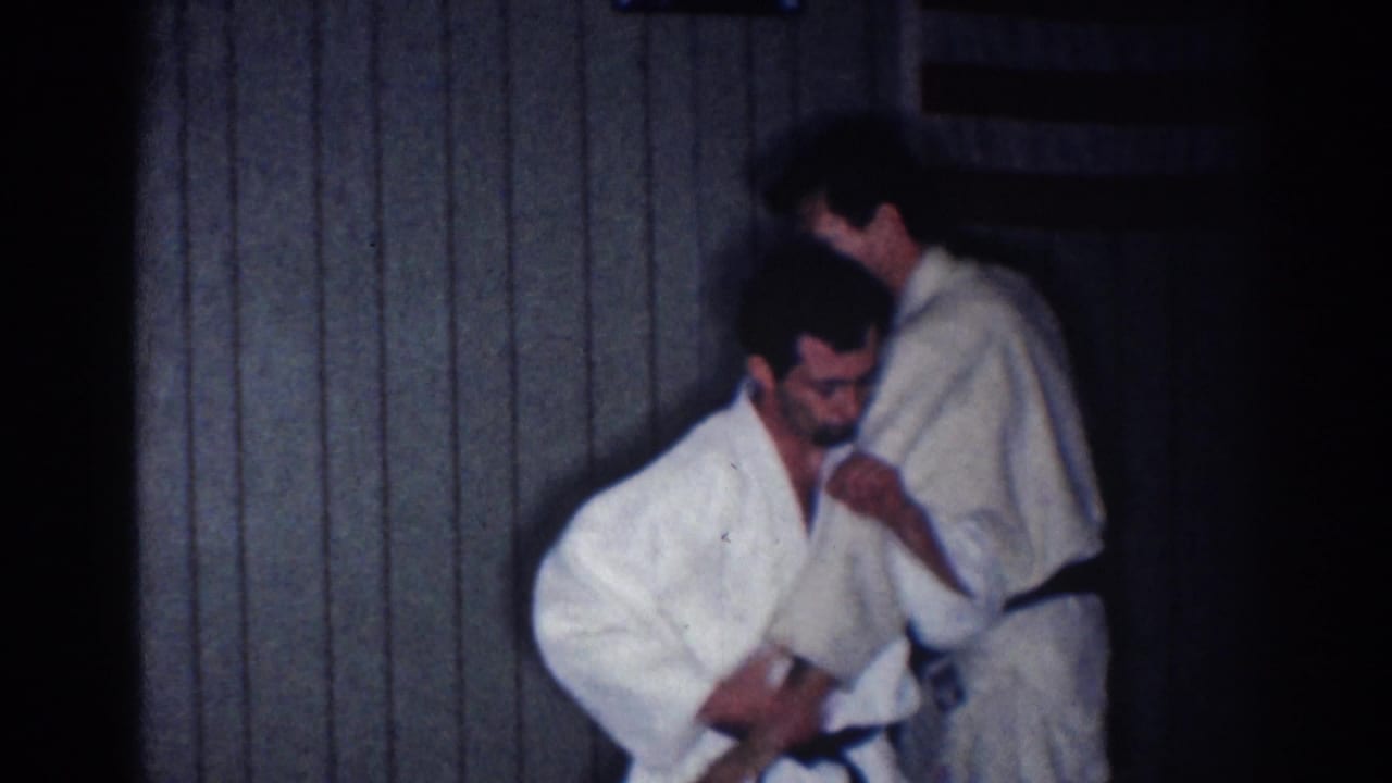 One practitioner crouches or dips forward while the other stands nearby in a dim 1967 Los Angeles indoor practice scene.