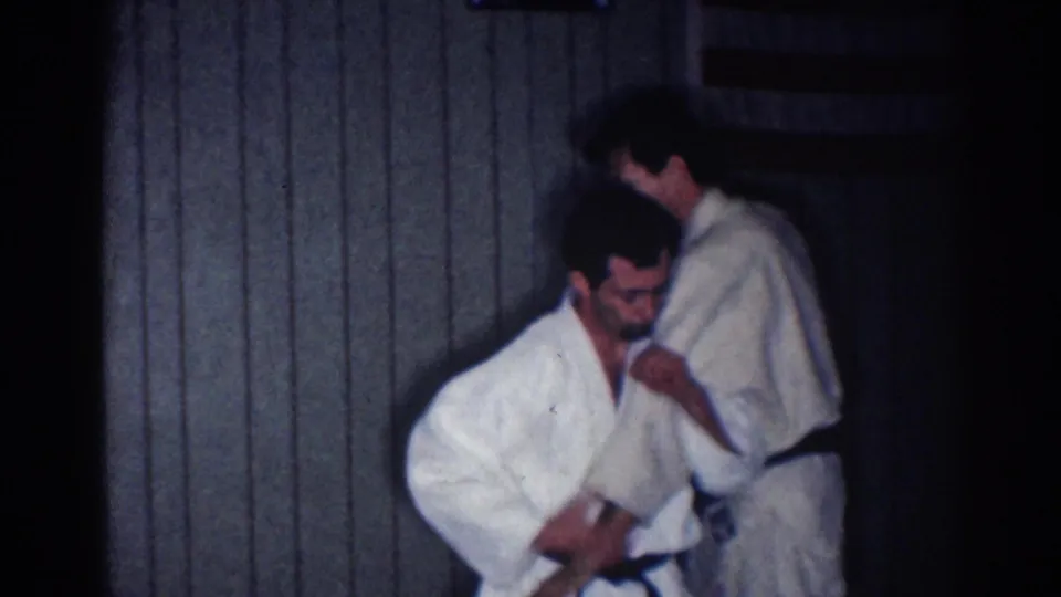 One practitioner crouches or dips forward while the other stands nearby in a dim 1967 Los Angeles indoor practice scene.