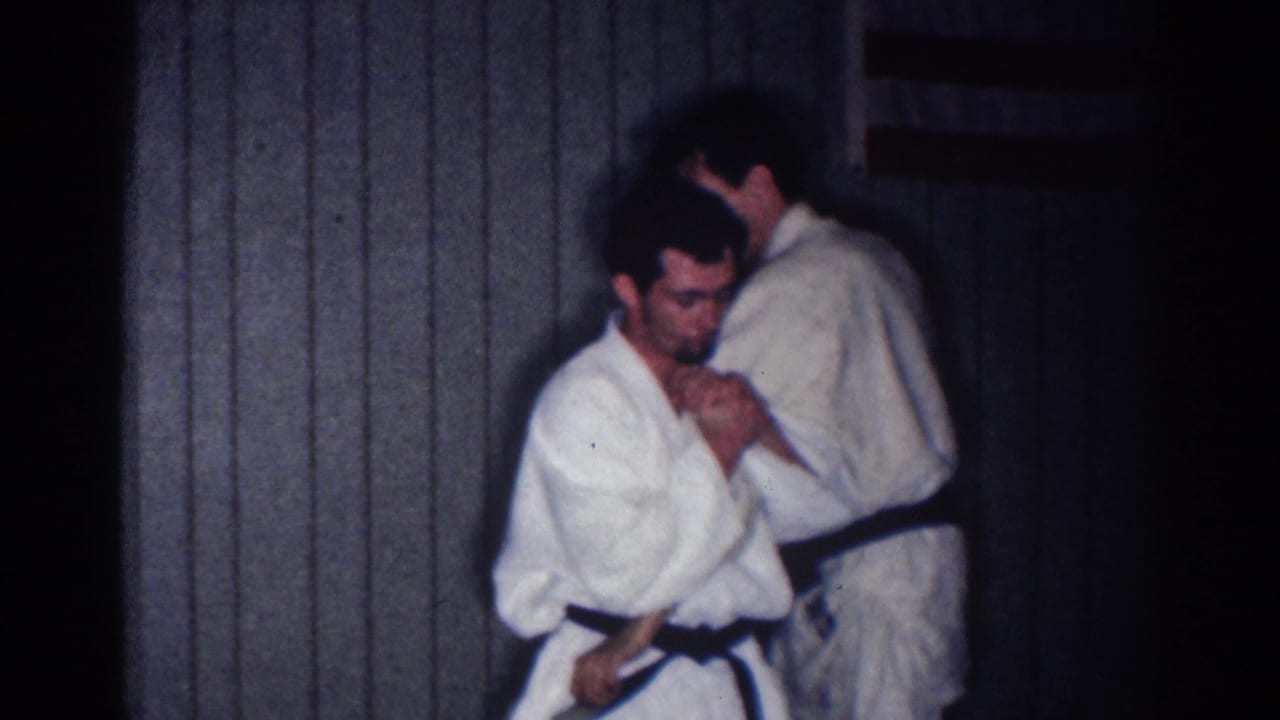 In a 1967 Los Angeles home-movie frame, two adult men in white judo gis prepare a close grappling hold indoors.