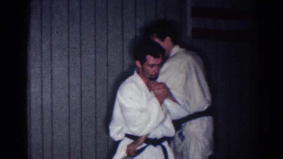 In a 1967 Los Angeles home-movie frame, two adult men in white judo gis prepare a close grappling hold indoors.