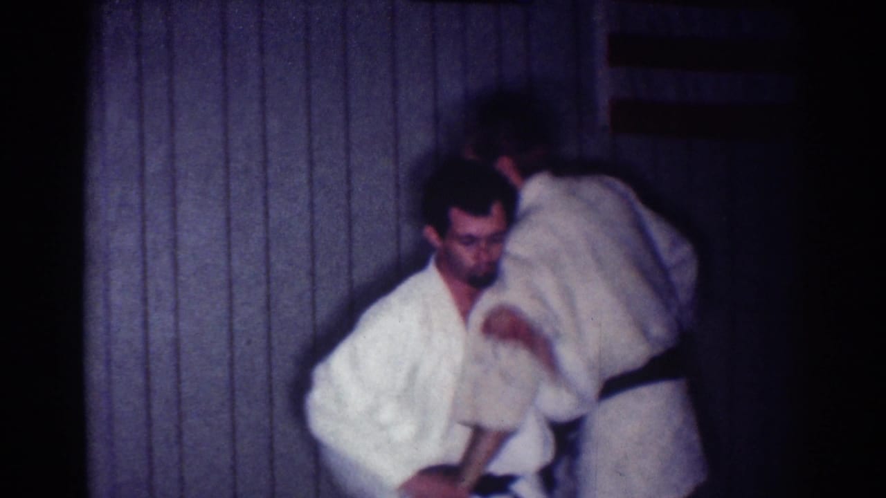 Two adult men in white gis demonstrate an indoor judo technique, with one partner bending as the other controls the motion.