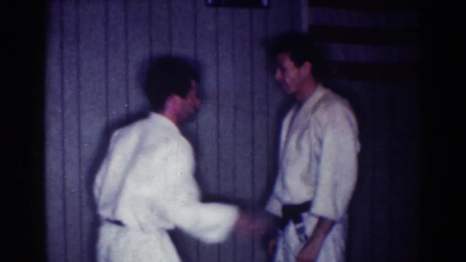 The partners face each other in the indoor 1967 judo practice footage, with motion blur from a quick arm movement.
