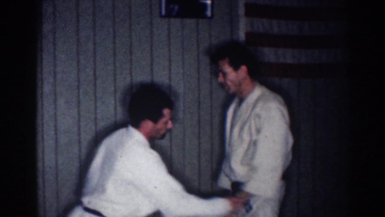 Two men in white gi practice a martial arts hold in a dim wood-paneled room.