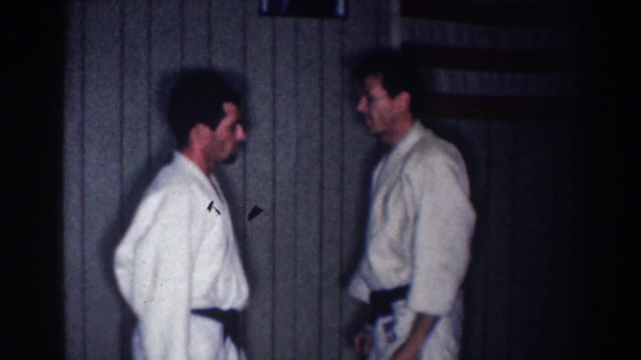 Two adult martial arts practitioners in white gi and black belts stand facing one another inside a Los Angeles training room.