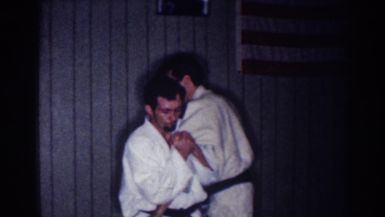 A man in a white gi faces the camera as another practitioner stands close behind during a martial arts drill indoors.