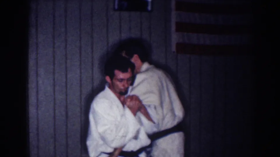 A man in a white gi faces the camera as another practitioner stands close behind during a martial arts drill indoors.