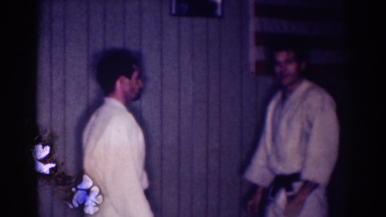Two men in white gi and black belts stand apart in a dim 1967 Los Angeles indoor martial arts practice space.