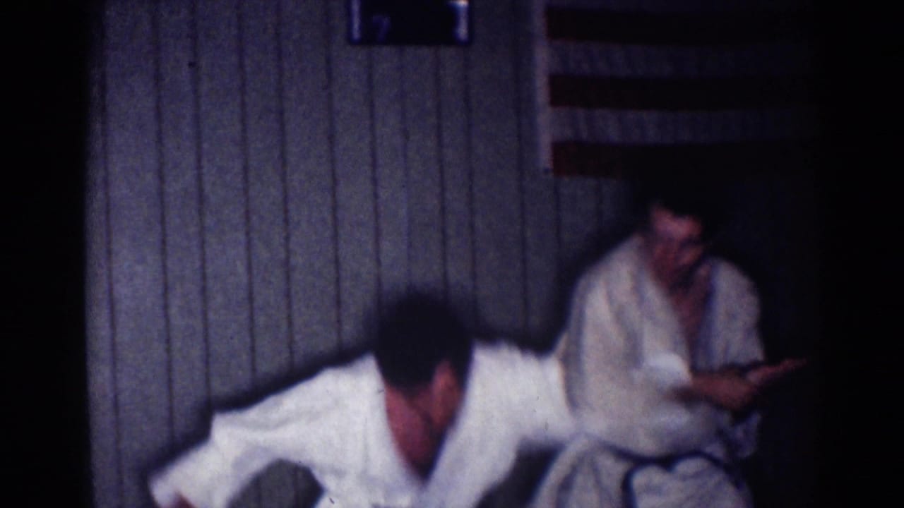 1967 Los Angeles 8mm frame showing two adult men in white karate gi during indoor practice near a wood-paneled wall.