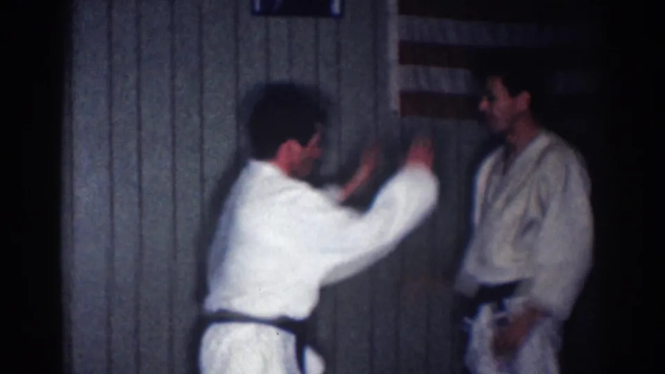 1967 Los Angeles karate practice frame with one man demonstrating a raised-arm movement toward another man in a white gi.