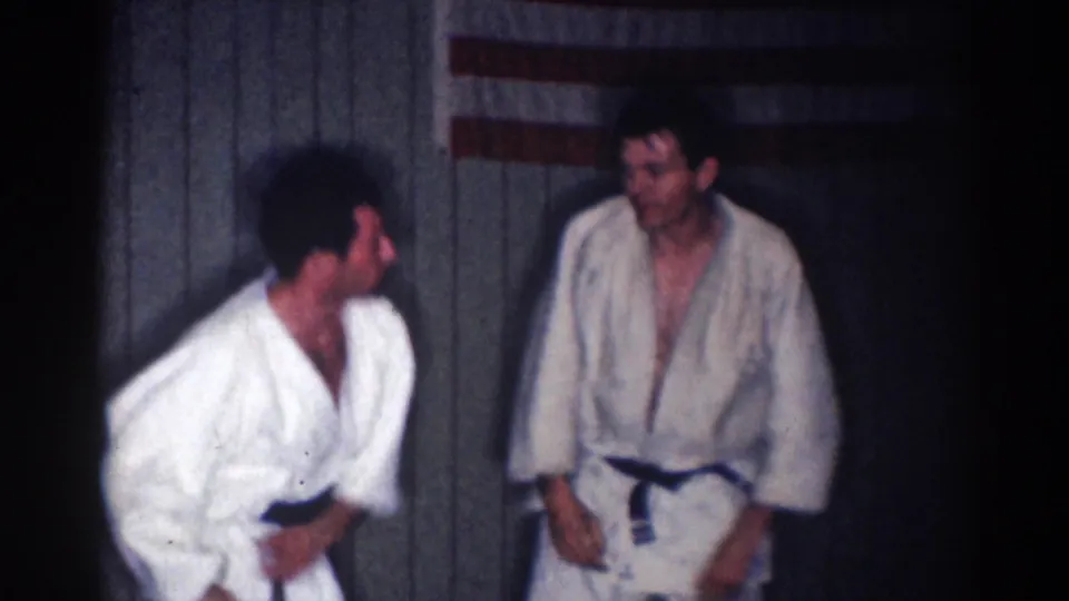 1967 Los Angeles 8mm still of two adult men in karate uniforms pausing or resetting during practice indoors.