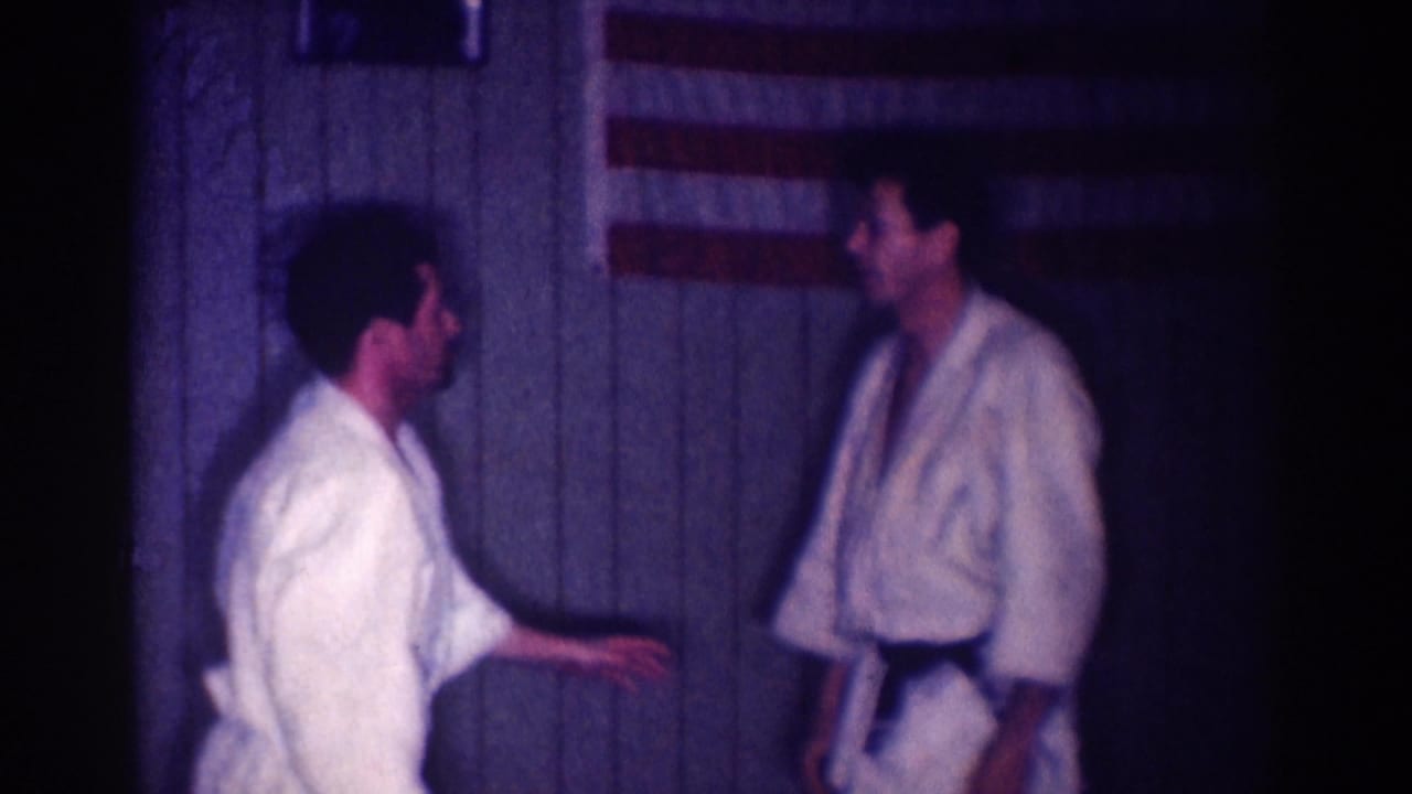 Two young men in white judo gis appear to practice in a dim Los Angeles interior, with an American flag on the paneled wall.