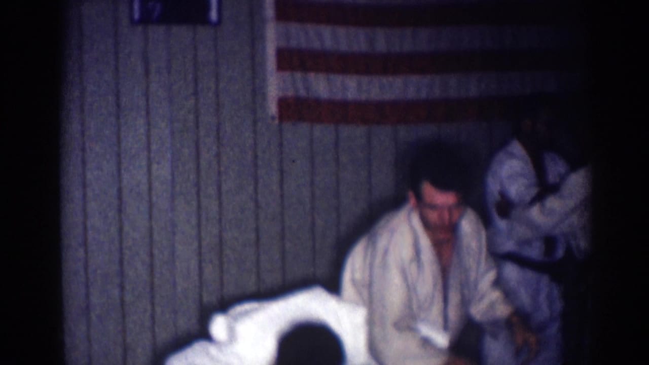 Judo practitioners move low to the floor near a paneled wall and flag.