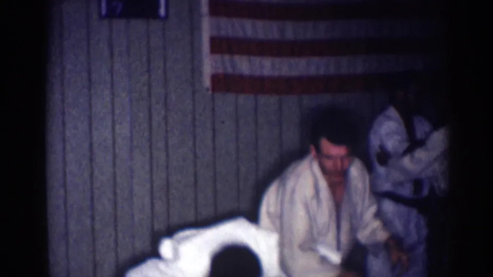 One judo practitioner crouches as another moves nearby in a dim 1967 home-movie frame from Los Angeles.