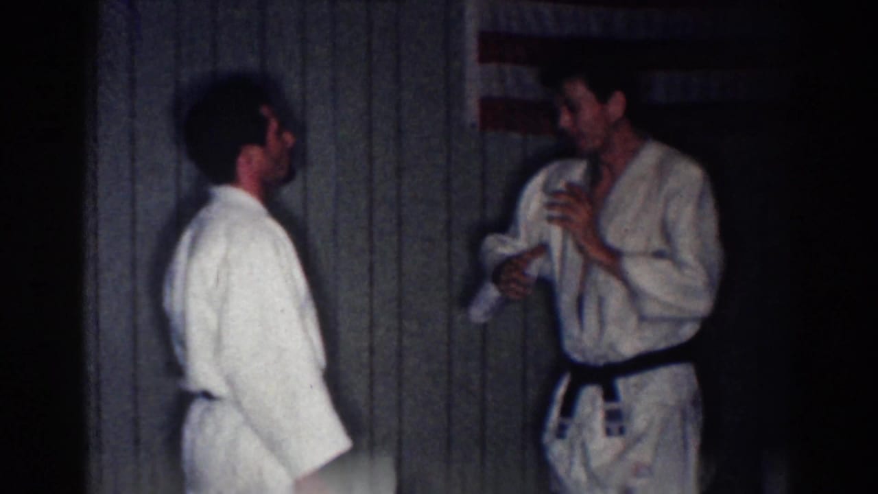 Two judo practitioners stand face to face in a dim private interior, one adjusting his gi near a dark belt.