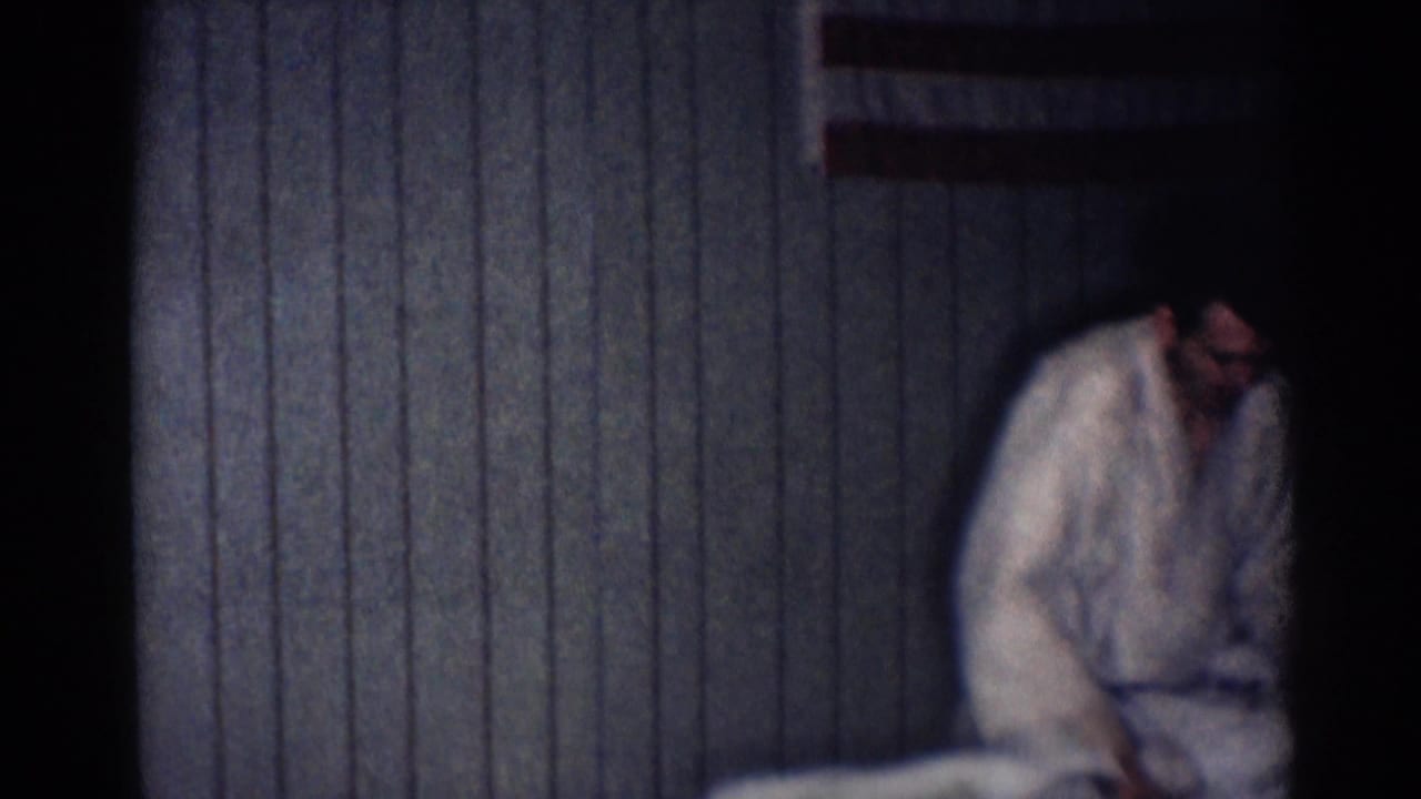 A man in a white judo gi bends toward the floor near the edge of frame in the dim Los Angeles training interior.