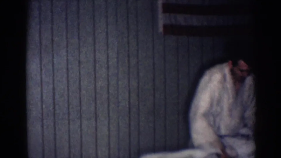 A man in a white judo gi bends toward the floor near the edge of frame in the dim Los Angeles training interior.