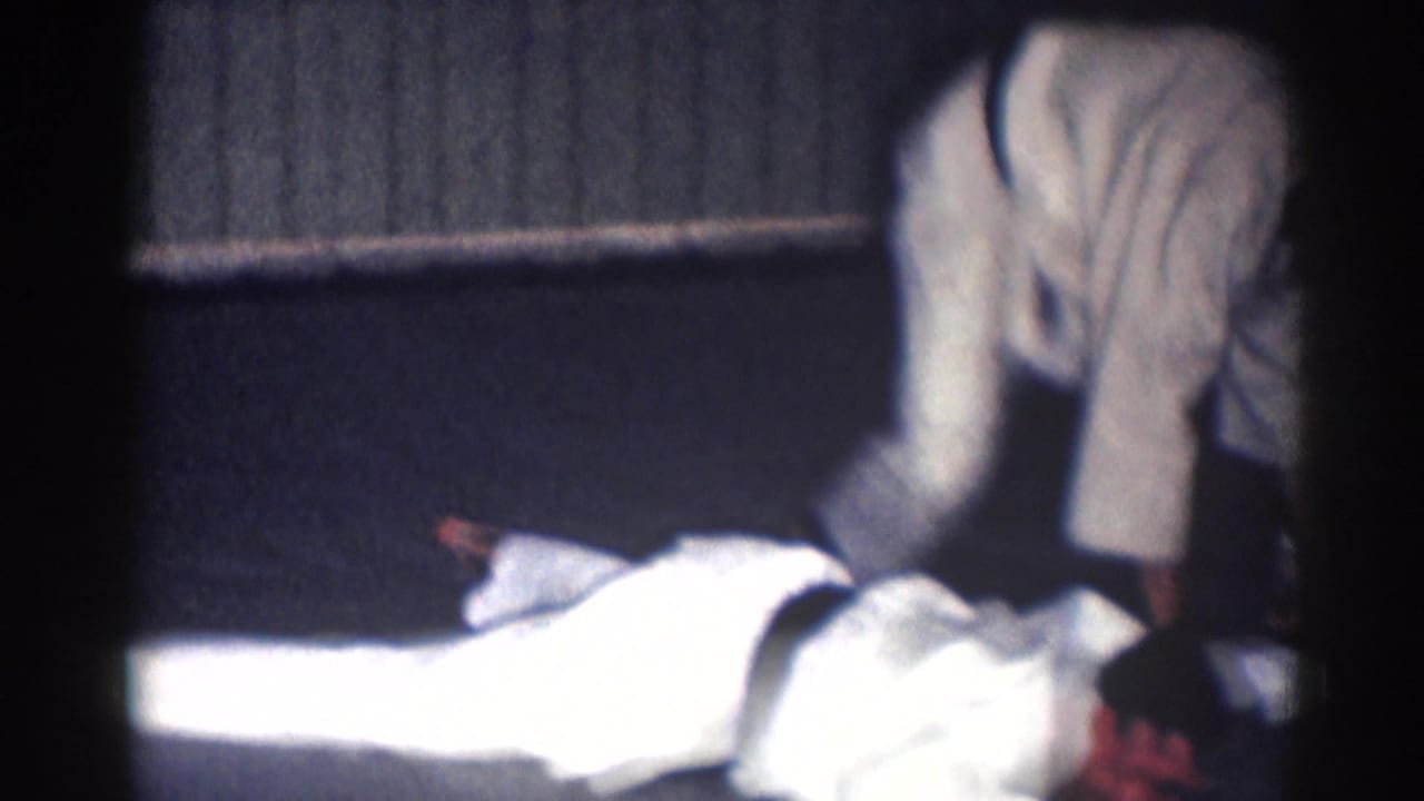 1967 Los Angeles 8mm frame showing two adult men in white gi during a low grappling or pinning technique indoors.