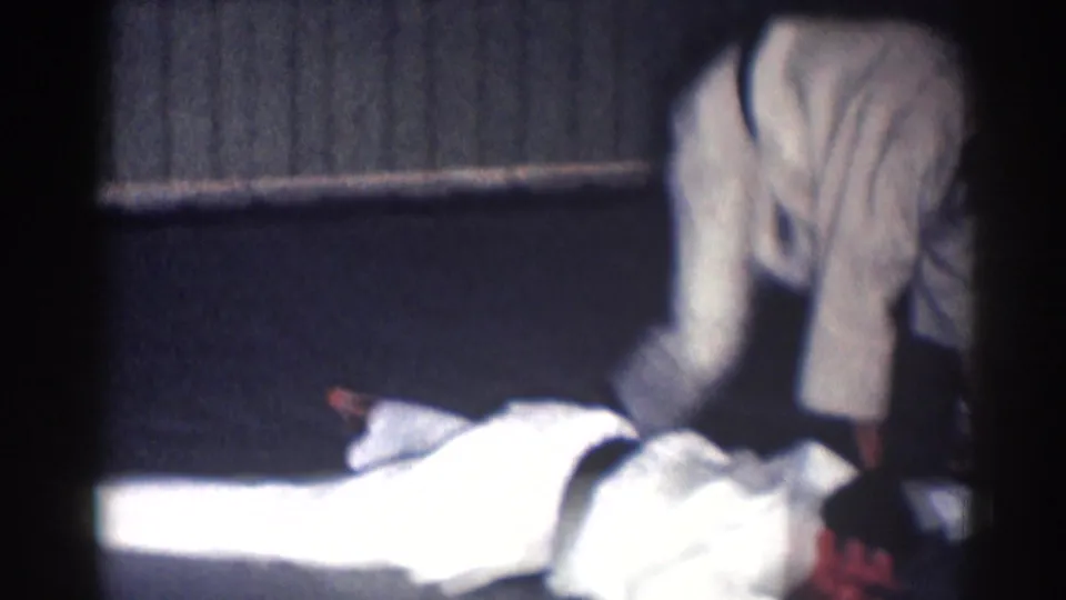 1967 Los Angeles 8mm frame showing two adult men in white gi during a low grappling or pinning technique indoors.