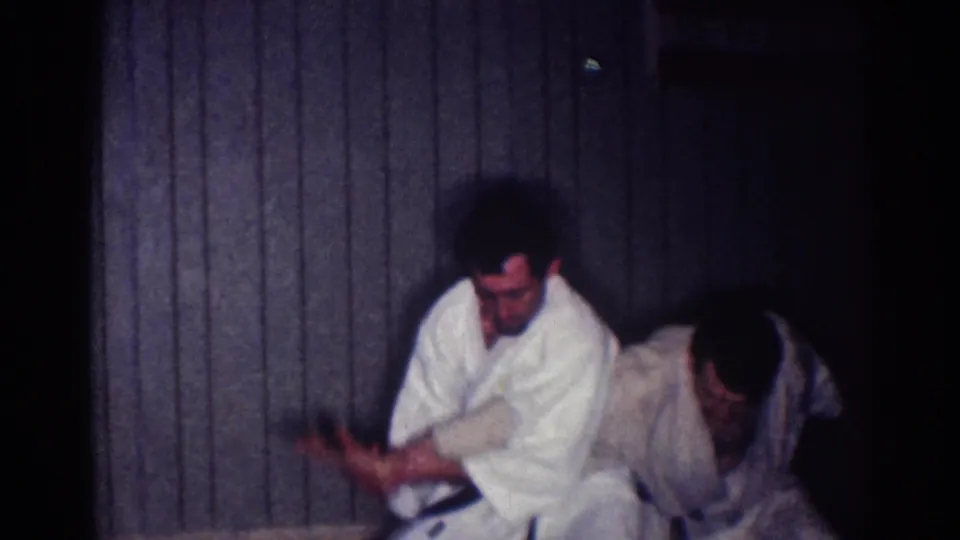 1967 Los Angeles 8mm frame showing two men in white gi working through a low armlock or hold during indoor practice.