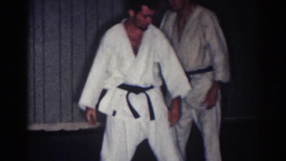 1967 Los Angeles frame showing two adult men in white gi standing near a paneled wall after a practice exchange.