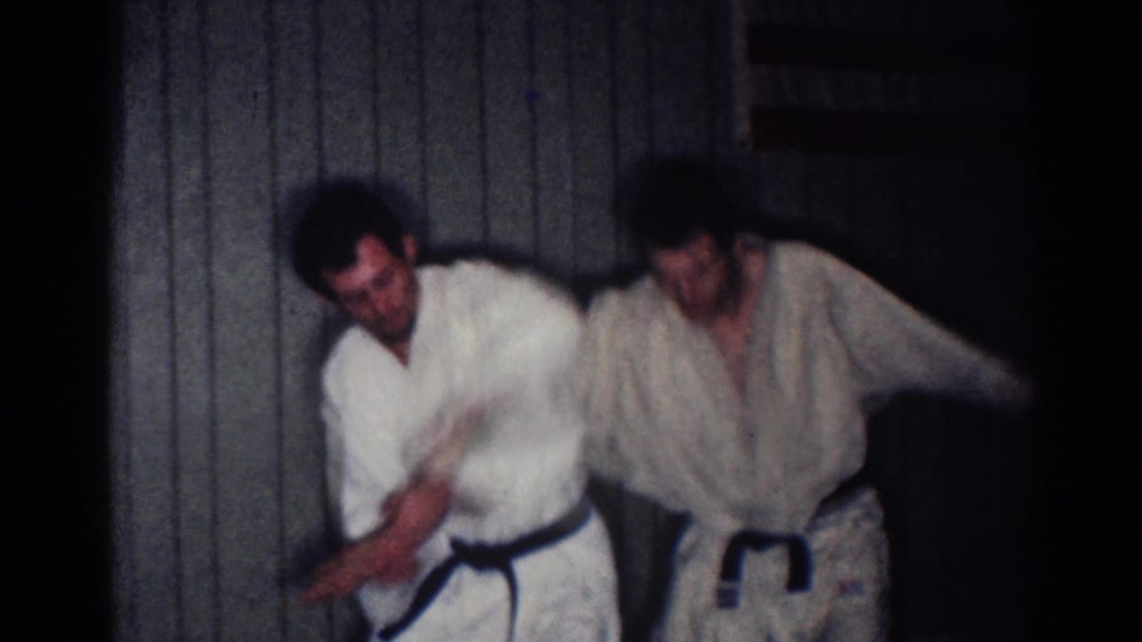 In 1967 Los Angeles footage, two adult men in white gi work through an arm-lock technique against a paneled wall.