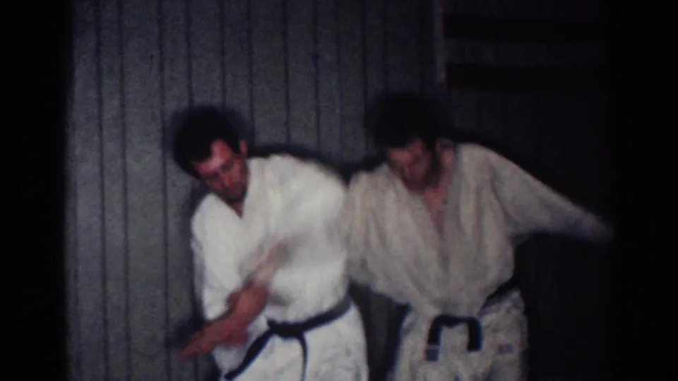 In 1967 Los Angeles footage, two adult men in white gi work through an arm-lock technique against a paneled wall.