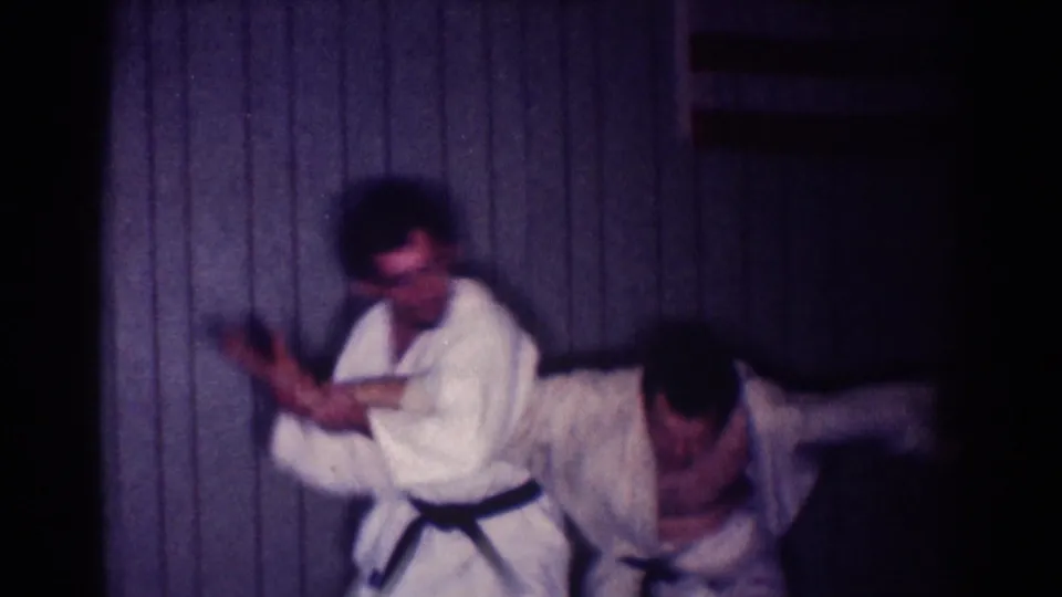 In a blurred 8mm frame, one man bends while the other applies an arm-control technique in an indoor Los Angeles practice setting.
