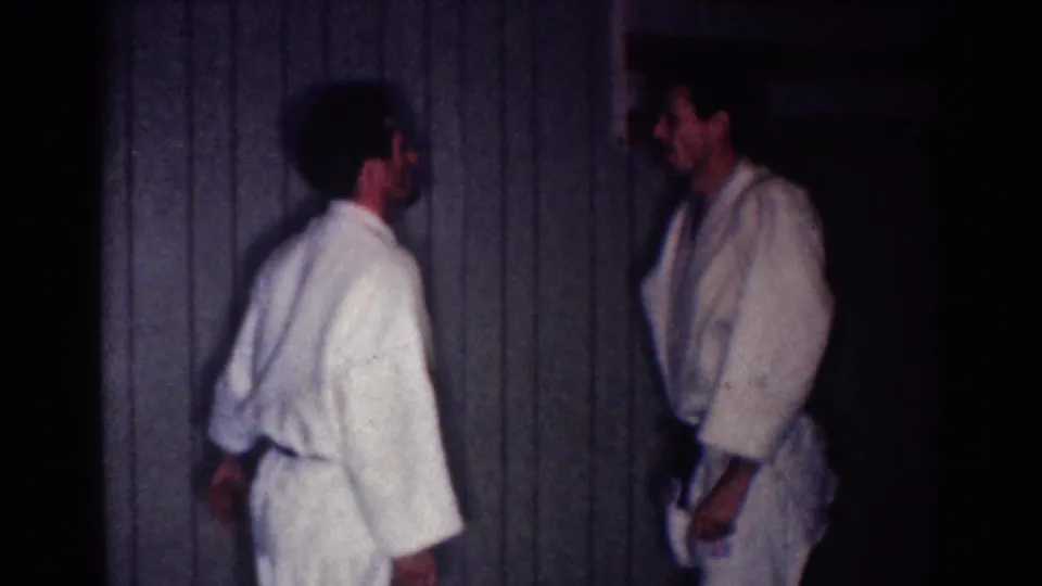 Two adult men in white gi stand facing one another in the dim paneled room during the 1967 Los Angeles karate practice footage.