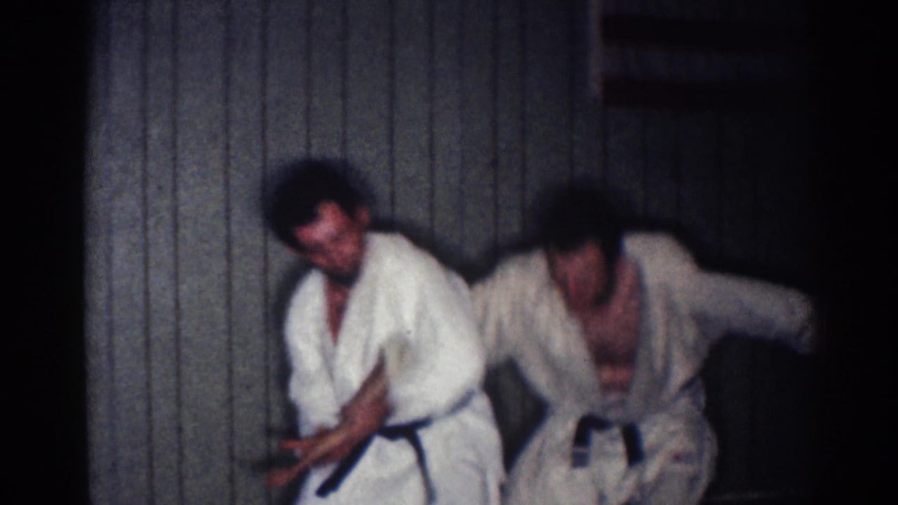 Two men in white karate gi practice a close-range block or strike indoors.