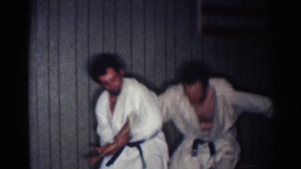 In 1967 Los Angeles home-movie footage, two adult karate students in white gi and dark belts practice a close-range technique against a paneled wall.