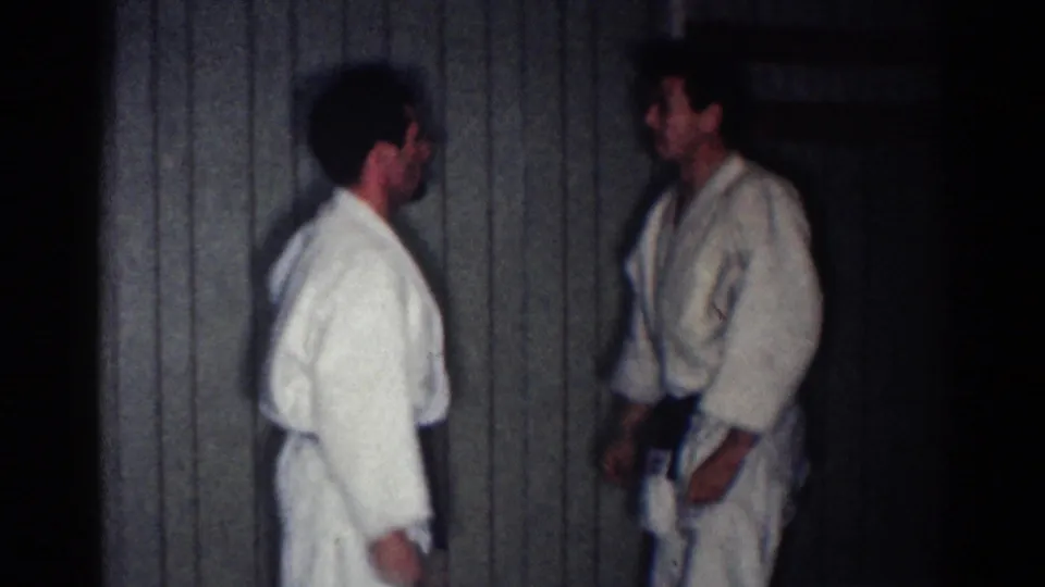Two adult karate students in white gi and dark belts face each other in a dim Los Angeles indoor practice space, consistent with the 1967 clip metadata.