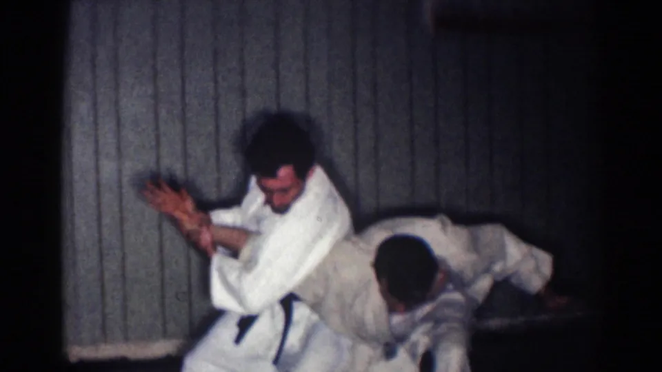 A karate student drops low during a throw or takedown practice while his partner raises his hands in the dim 1967 Los Angeles home-movie scene.