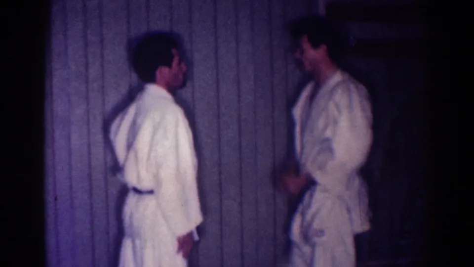 Two adult karate students in white gi face each other near a paneled wall, with a purple color shift in the 1967 Los Angeles 8mm footage.