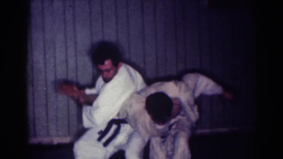 Two adult men in white gi and dark belts practice a close sparring move in 1967 Los Angeles home-movie footage.