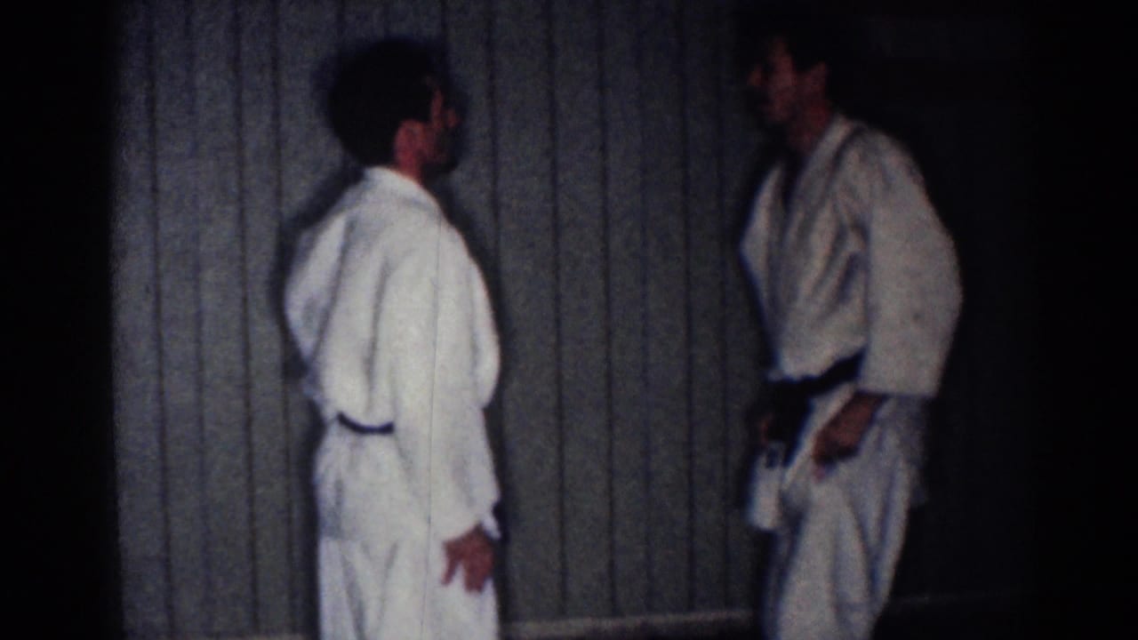 Two men in white gi stand facing each other beside a paneled wall.