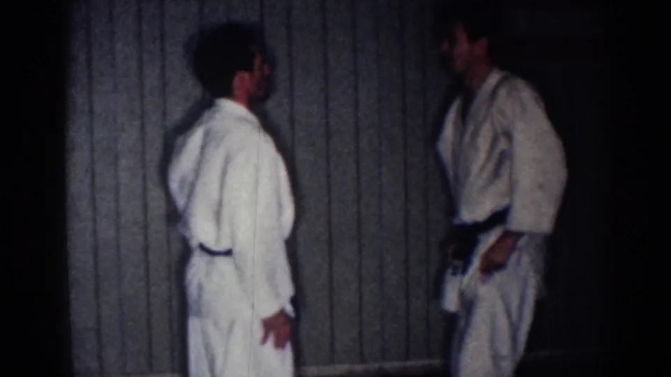 Two adult martial arts practitioners in white gi face each other in a dim interior with vertical wood paneling.