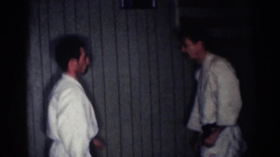 Two adult practitioners in white martial arts uniforms stand facing each other in a low-lit 1967 Los Angeles home movie.