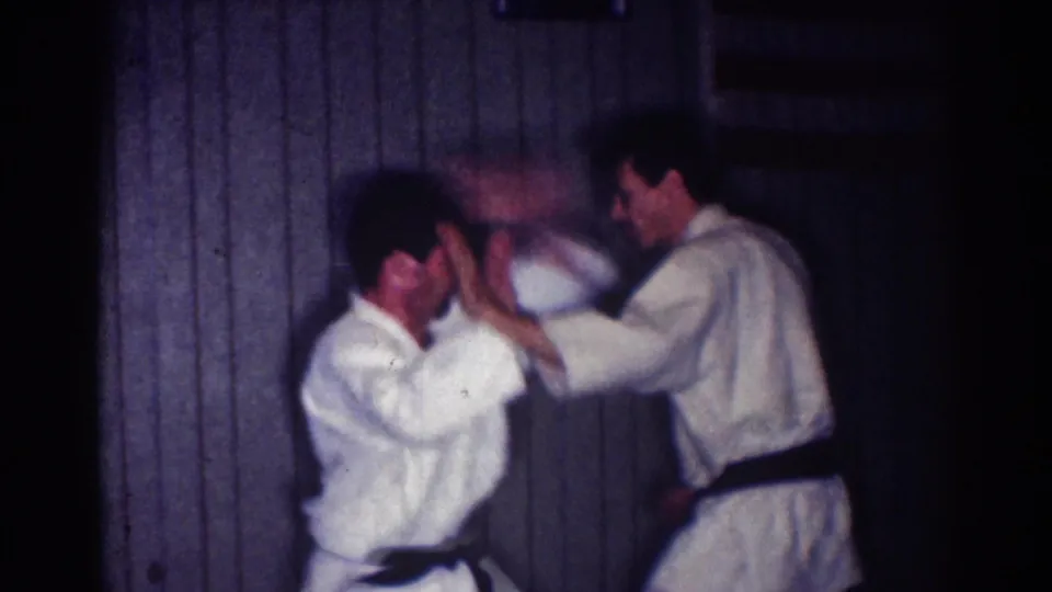In 1967 Los Angeles home-movie footage, two adult men in white judo gis and black belts practice a close-range defensive movement indoors.