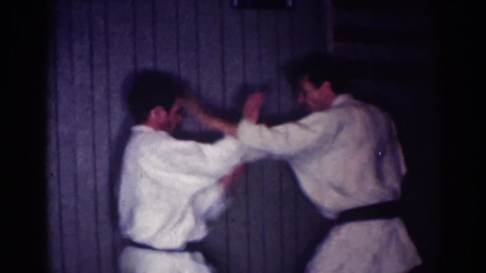 Two adult men in white gis and black belts face each other in a dim Los Angeles interior, arms lifted during a judo practice exchange.
