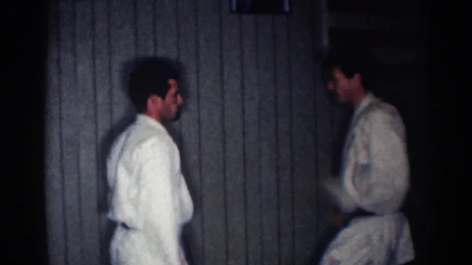 The practitioners pause face-to-face in white judo uniforms and black belts, filmed indoors on 8mm home-movie footage in Los Angeles.