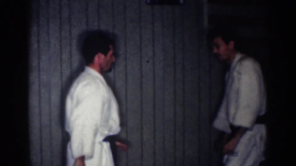 Two adult black-belt judo practitioners stand apart in a dim paneled interior, one smiling or relaxed after the practice exchange.
