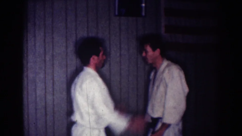 Two adult men in white gi practice a close-range karate movement in the dim Los Angeles interior noted in clip metadata.