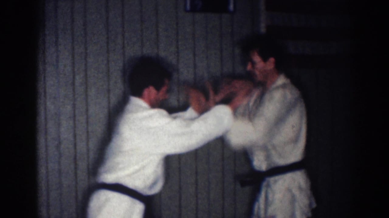 Two men in white karate gis spar indoors, one extending his hands toward the other.