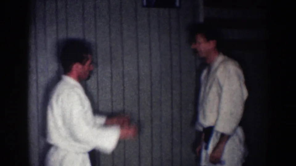 Two adult men in white gis and black belts stand facing each other during a paused sparring moment in a Los Angeles interior.