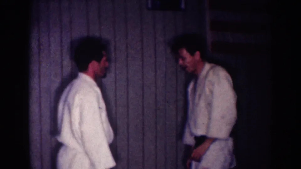 Two adult men in white karate gis stand in a dim wood-paneled room, appearing to reset after a sparring exchange.